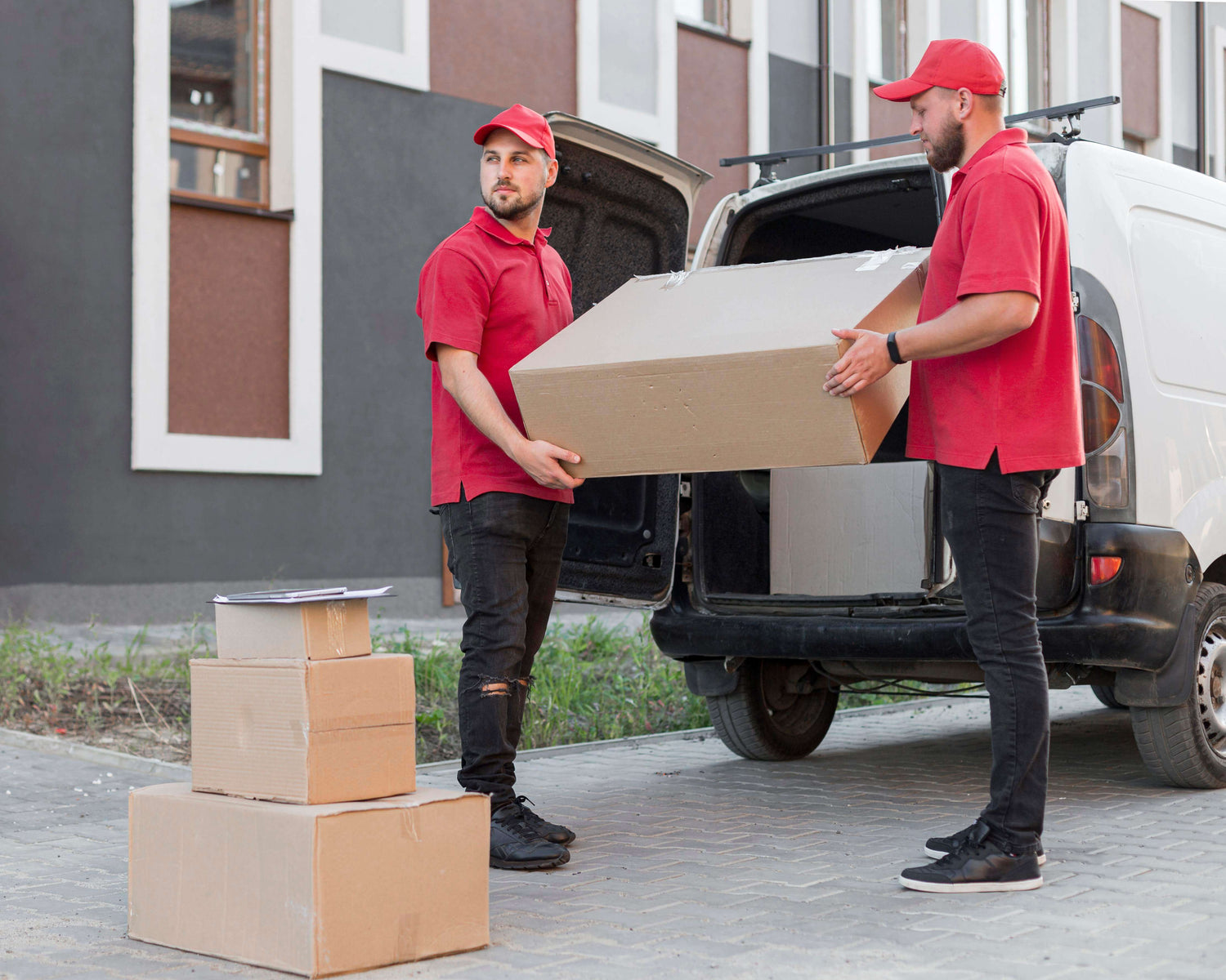 Two delivery workers in red shirts unloading boxes from a van outside a building. 24BIZHUB